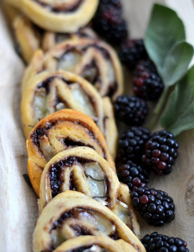 Brie and blackberry jam pinwheels on a wooden tray adorned with fresh blackberries and sage leaves.