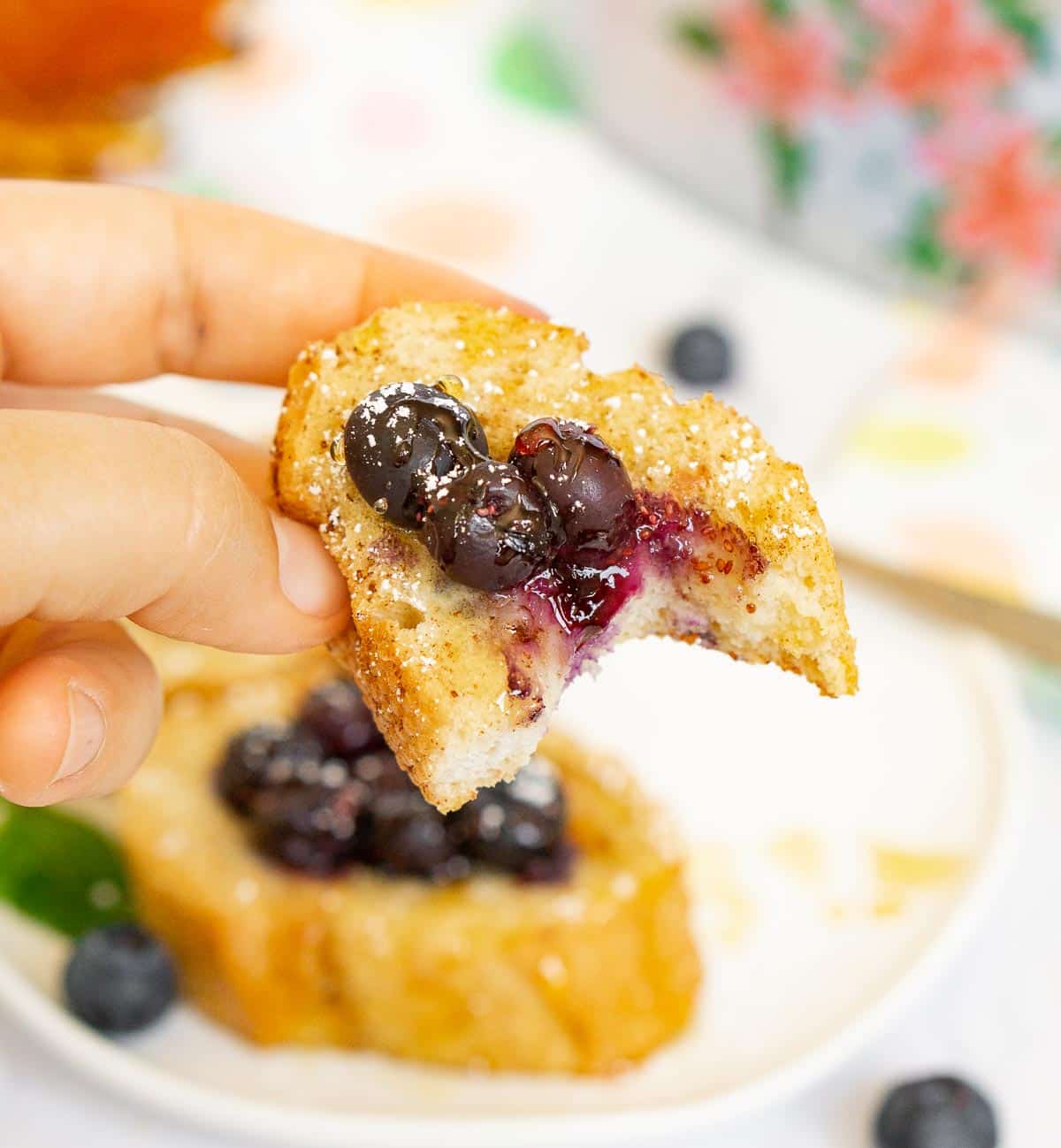 Hand holding piece of baguette French toast with blueberries with a bite taken out to show texture