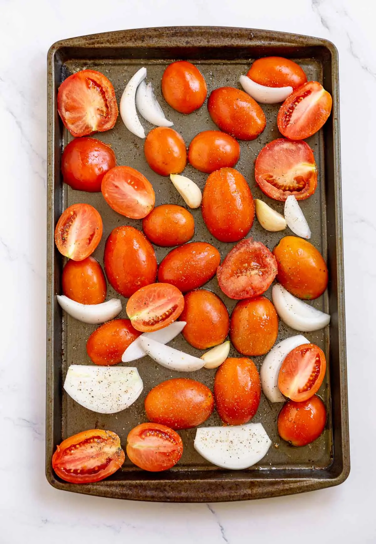 Halved tomatoes, sliced onions, and garlic cloves on a roasting sheet