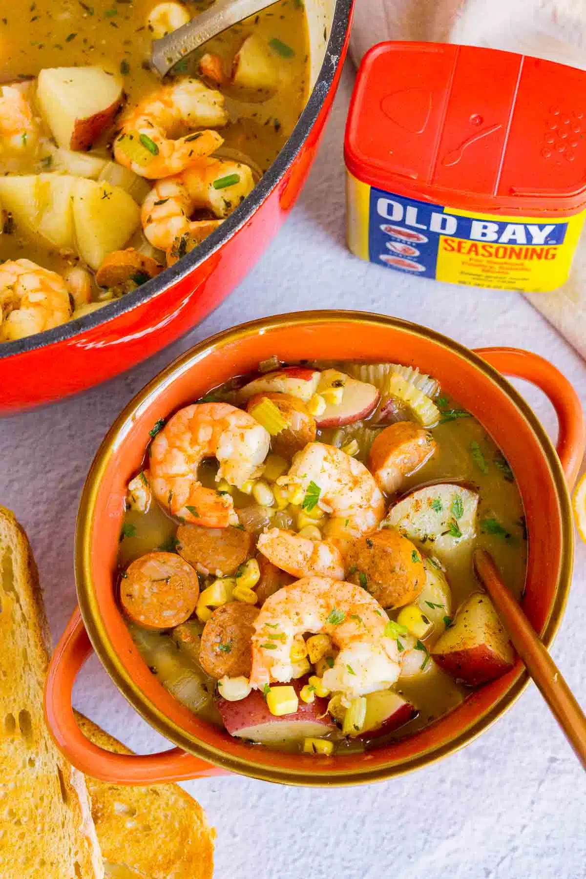 Shrimp boil soup in a bowl with crusty bread and Old Bay nearby